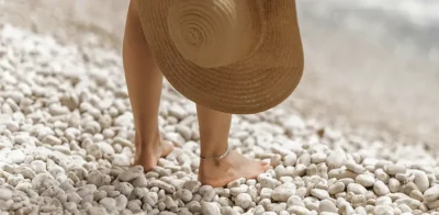En kvinde står barfodet på en stenet strand med en hat i hånden.