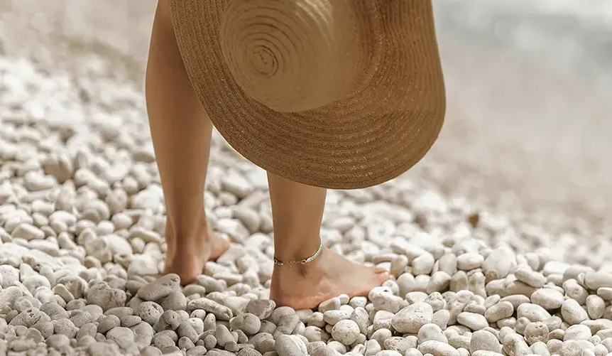 Una mujer de pie, descalza, en una playa de piedras, con un sombrero en la mano.