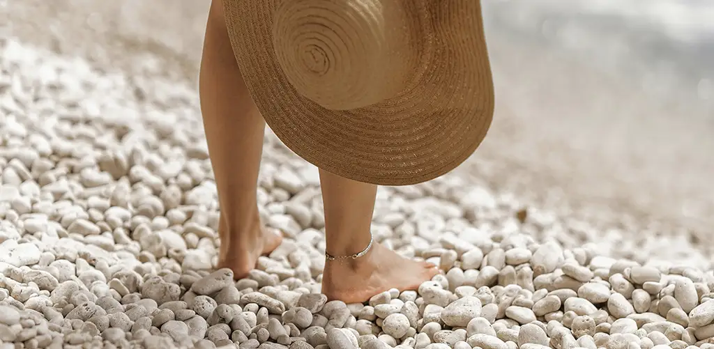 A woman standing barefoot on a stony beach, holding a hat.