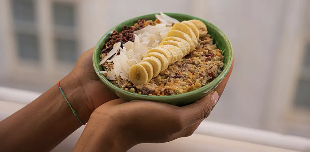 mains tenant un bol vert avec des aliments sains, des flocons de coco, des bananes et des graines.