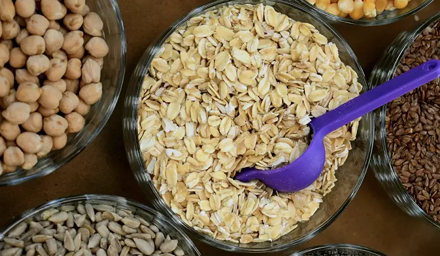 Primer plano de avena, garbanzos y semillas en cuencos pequeños, con una cucharita de plástico morada metida en la avena