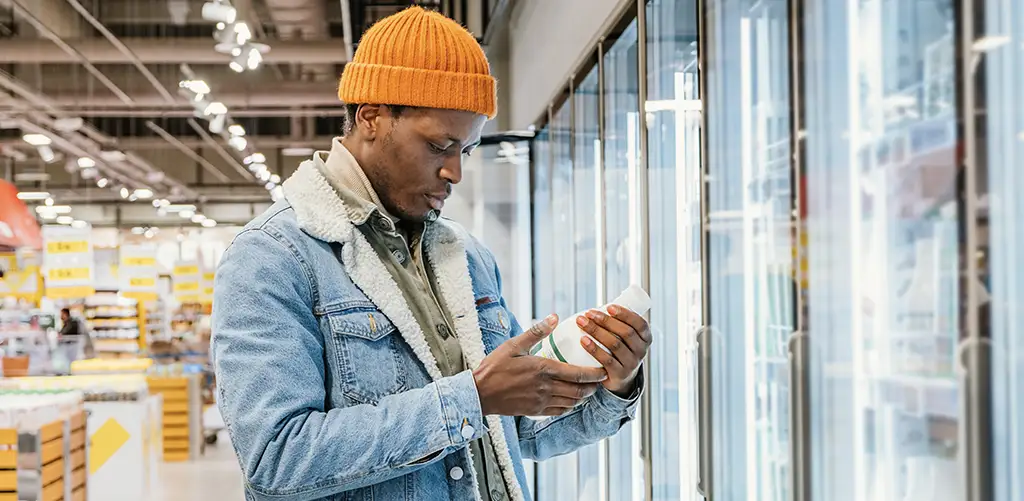 Un homme portant un bonnet orange et une veste en jean lit l'étiquette d'un produit alors qu'il fait ses courses dans le rayon réfrigéré d'une épicerie.