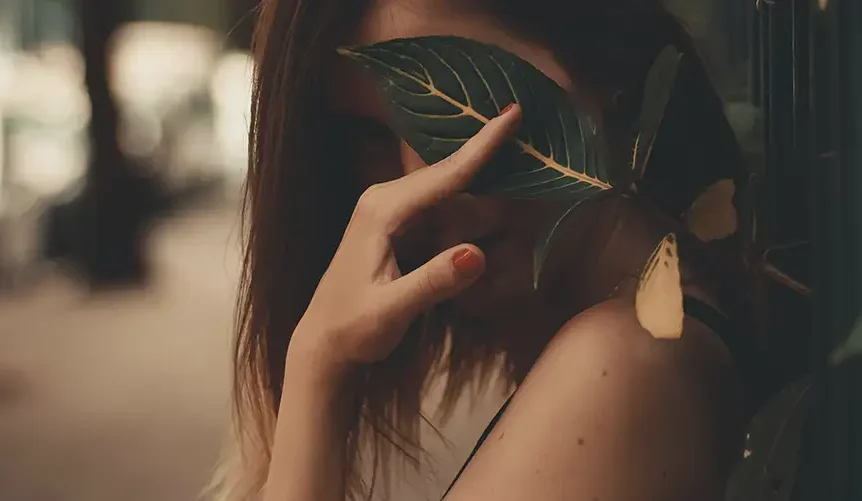 brown-haired woman with red coloured nails hide her face with a leave