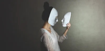 Eine Frau wird im Profil mit einer weißen Maske fotografiert, während sie eine identische weiße Maske in den Händen hält und sie anschaut. Sie trägt ein geschnürtes weißes Kleid.