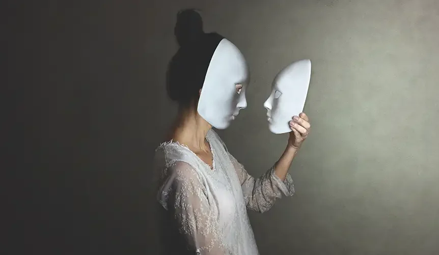 Une femme est photographiée de profil, portant un masque blanc, tout en tenant dans ses mains un masque blanc identique qu'elle observe. Elle porte une robe blanche en dentelle.