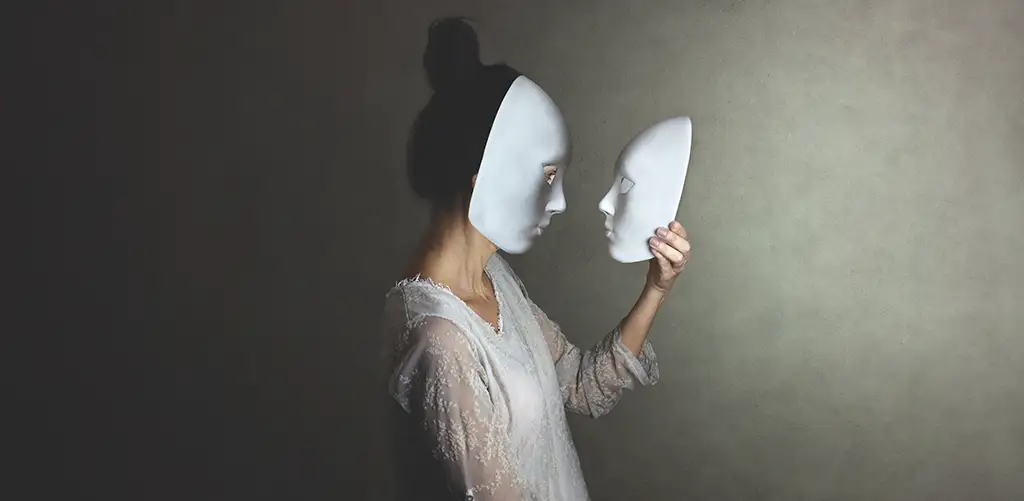 A woman is photographed in profile wearing a white mask, while holding an identical white mask in her hands and looking at it. She is wearing a laced white dress.