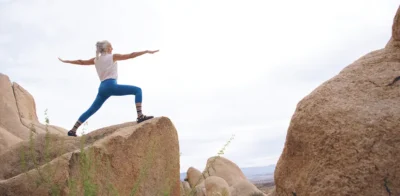 An older woman wearing blue tights practices yoga in a rocky coastal landscape with a clear view of the sea. She holds a balanced pose with calm focus, surrounded by cliffs and open sky.