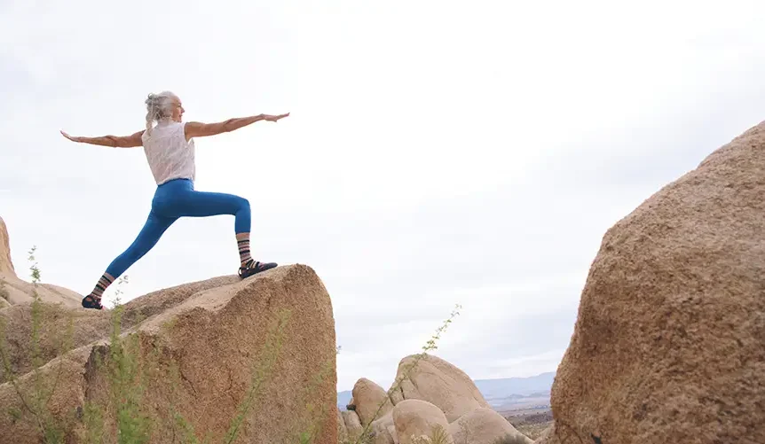 An older woman wearing blue tights practices yoga in a rocky coastal landscape with a clear view of the sea. She holds a balanced pose with calm focus, surrounded by cliffs and open sky.