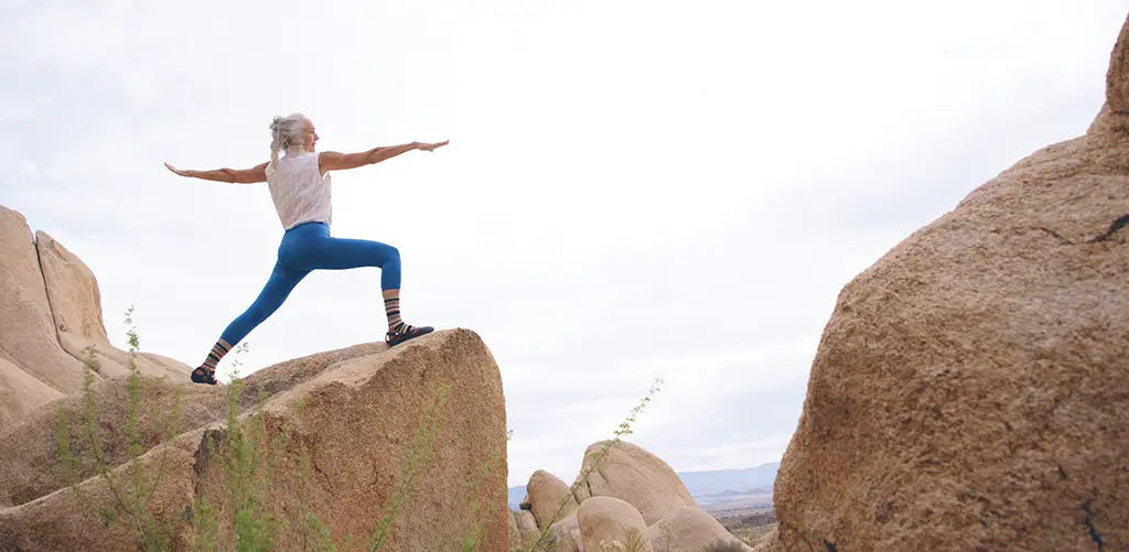 O femeie în vârstă purtând colanți albaștri practică yoga într-un peisaj stâncos de coastă cu vedere clară la mare. Ea ține o poziție echilibrată cu o concentrare calmă, înconjurată de stânci și cer deschis.