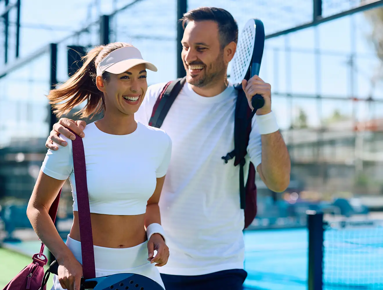 Un homme et une femme souriants marchant ensemble sur un terrain de padel en plein air, tenant des raquettes de padel et portant du matériel de sport.