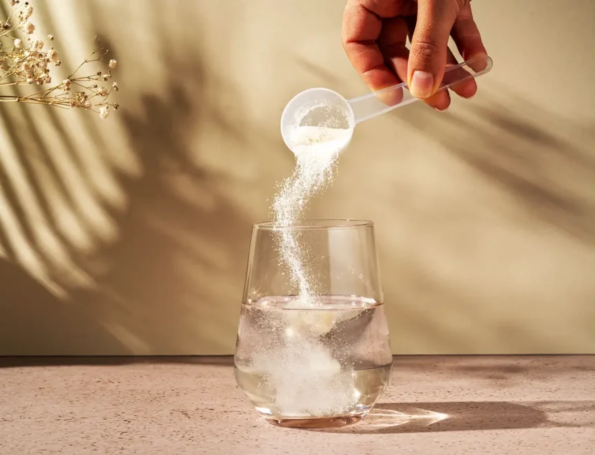 Je giet met de hand een schepje proteïnepoeder in een glas met een gele drank, terwijl er nog een schepje poeder naast het glas op een wit oppervlak ligt.