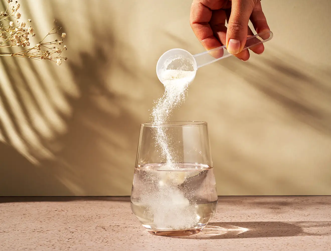 Se ve cómo se echa a mano una cucharada de proteína en polvo en un vaso de una bebida amarilla, con otra cucharada de proteína en polvo colocada junto al vaso sobre una superficie blanca.