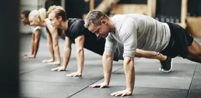 A group of people exercising together in a gym, doing push-ups on the floor. The focus is on a man in a gray workout shirt at the front, with other participants lined up behind him.