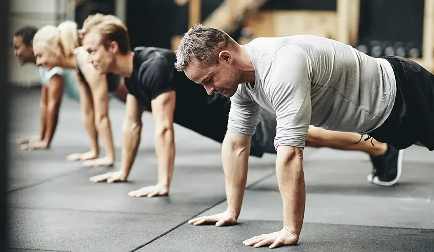 A group of people exercising together in a gym, doing push-ups on the floor. The focus is on a man in a gray workout shirt at the front, with other participants lined up behind him.