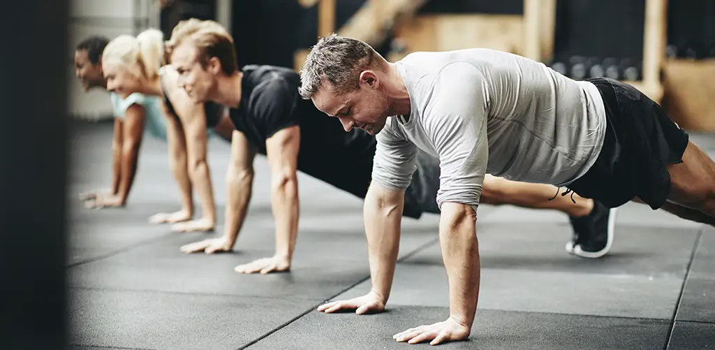 A group of people exercising together in a gym, doing push-ups on the floor. The focus is on a man in a gray workout shirt at the front, with other participants lined up behind him.