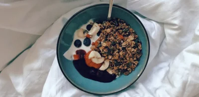 A bowl of yogurt topped with granola, banana slices, blueberries, and fruit jam, served with a spoon on a white bed sheet.