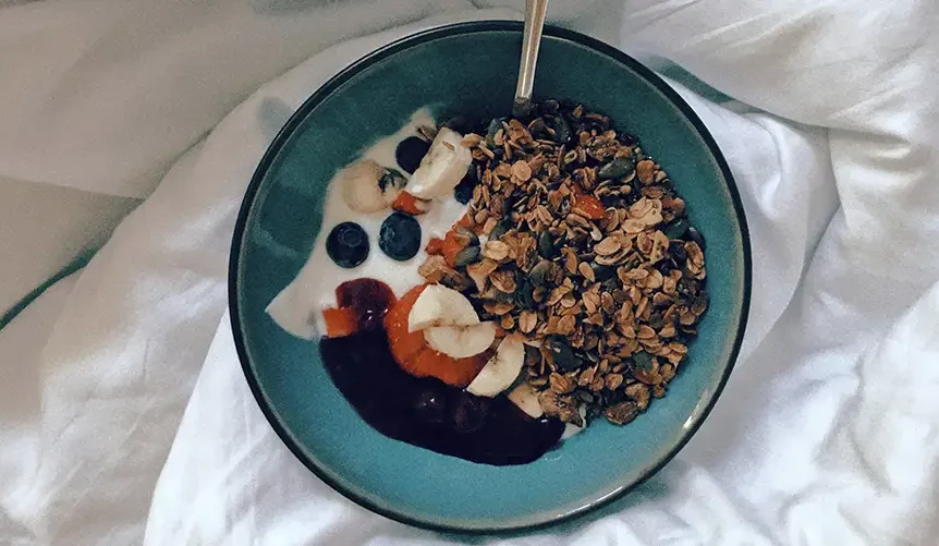 A bowl of yogurt topped with granola, banana slices, blueberries, and fruit jam, served with a spoon on a white bed sheet.