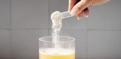 Einen Messlöffel Proteinpulver mit der Hand in ein Glas mit gelbem Getränk schütten und einen weiteren Messlöffel Pulver neben das Glas auf eine weiße Fläche legen.