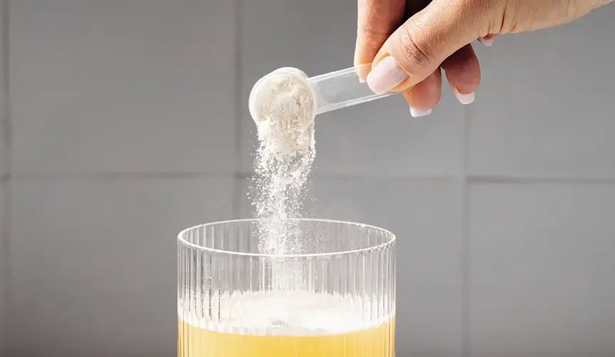 Hand pouring a scoop of protein powder into a glass of yellow drink, with another scoop of powder placed next to the glass on a white surface.