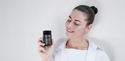 Una mujer sonriente con el pelo oscuro recogido en un moño, vestida con una camisa blanca, sostiene una botella negra de Purovitalis Berberine Balance sobre un fondo claro.