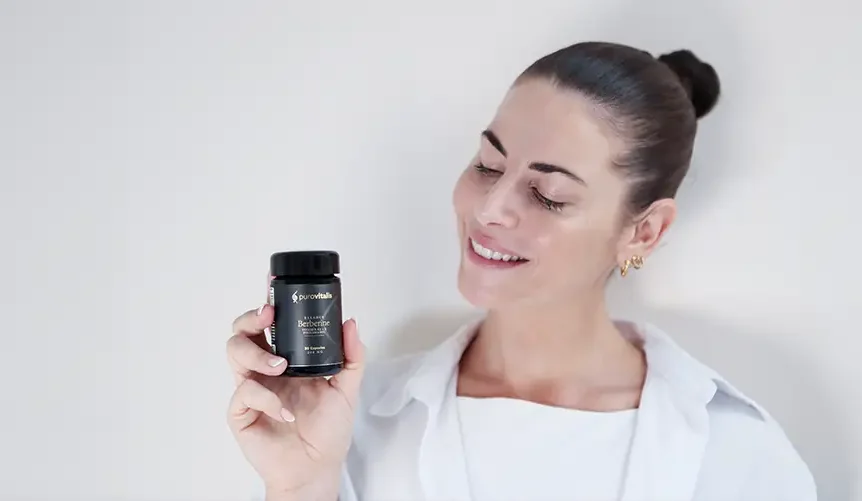 Una mujer sonriente, con el pelo oscuro recogido en un moño y vestida con una camisa blanca, sostiene un frasco negro de «Purovitalis Berberine Balance» sobre un fondo claro.