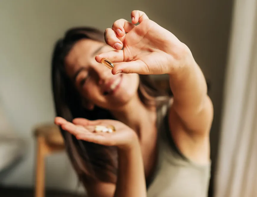 Een glimlachende vrouw met voedingssupplementen in haar hand, die een capsule tussen haar vingers laat zien, als symbool voor dagelijkse gezondheid en welzijn.