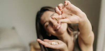 Een lachende vrouw houdt voedingssupplementen in haar hand, met een capsule tussen haar vingers, symbool voor dagelijkse gezondheid en welzijn.