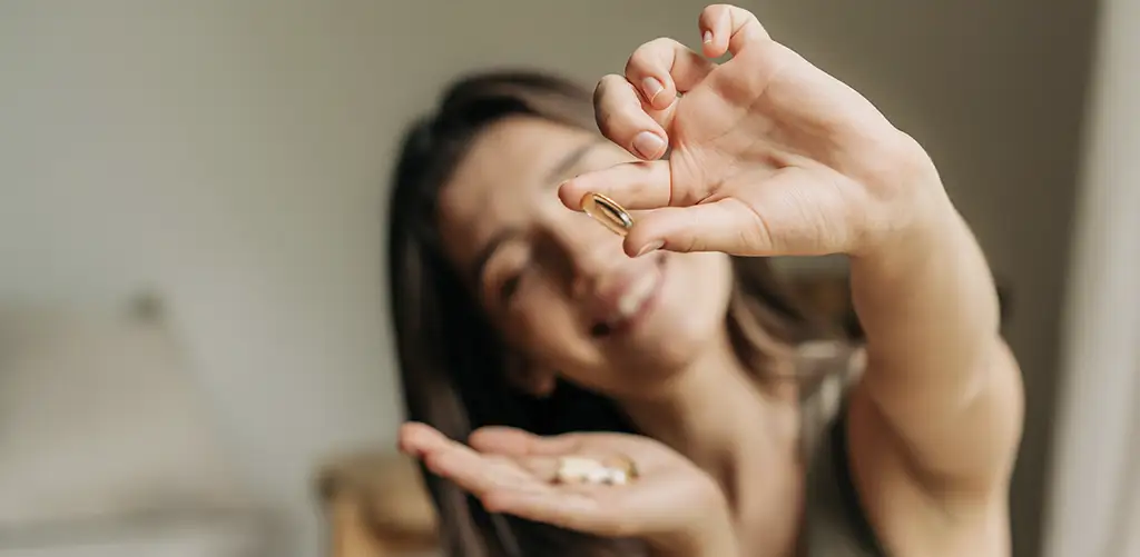 Een lachende vrouw houdt voedingssupplementen in haar hand, met een capsule tussen haar vingers, symbool voor dagelijkse gezondheid en welzijn.