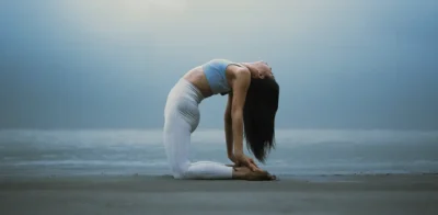 Une femme pratiquant le yoga dans la pose du chameau (Ustrasana) sur une plage à l'aube, vêtue d'un haut bleu clair et d'un legging blanc.