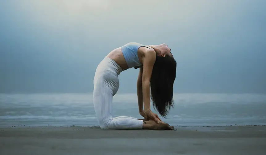 A woman practicing yoga in the camel pose (Ustrasana) on a beach at dawn, wearing a light blue top and white leggings