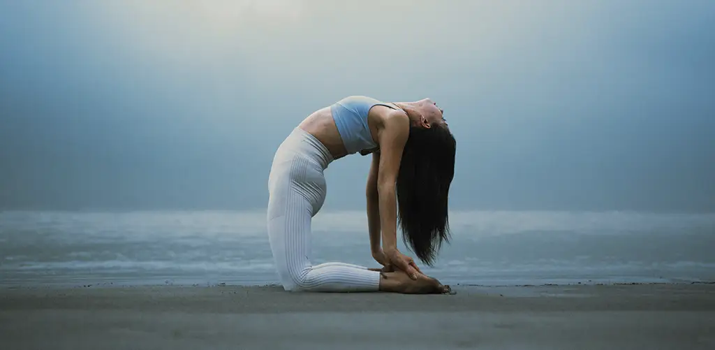 Une femme pratiquant le yoga dans la pose du chameau (Ustrasana) sur une plage à l'aube, vêtue d'un haut bleu clair et d'un legging blanc.