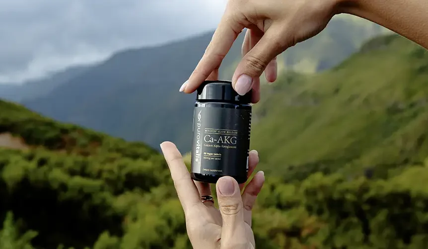 Hands holding a Purovitalis Ca-AKG supplement bottle outdoors with green mountains in the background.