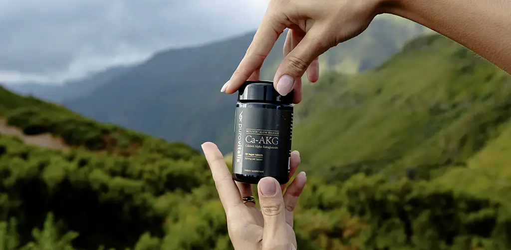 Hände halten eine Purovitalis Ca-AKG Ergänzungsflasche im Freien mit grünen Bergen im Hintergrund.