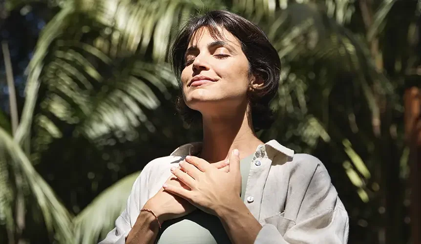 Una mujer está de pie al aire libre, con los ojos cerrados y una sonrisa serena, apoyando las manos en el pecho mientras respira profundamente. La luz del sol se cuela entre la vegetación tropical que la rodea, creando un ambiente tranquilo y de plenitud.