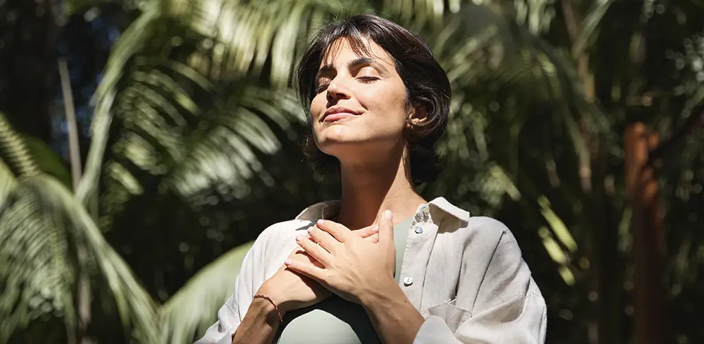 A woman stands outdoors with her eyes closed and a calm smile, resting her hands on her chest while breathing deeply. Sunlight filters through tropical greenery around her, creating a peaceful and mindful atmosphere.