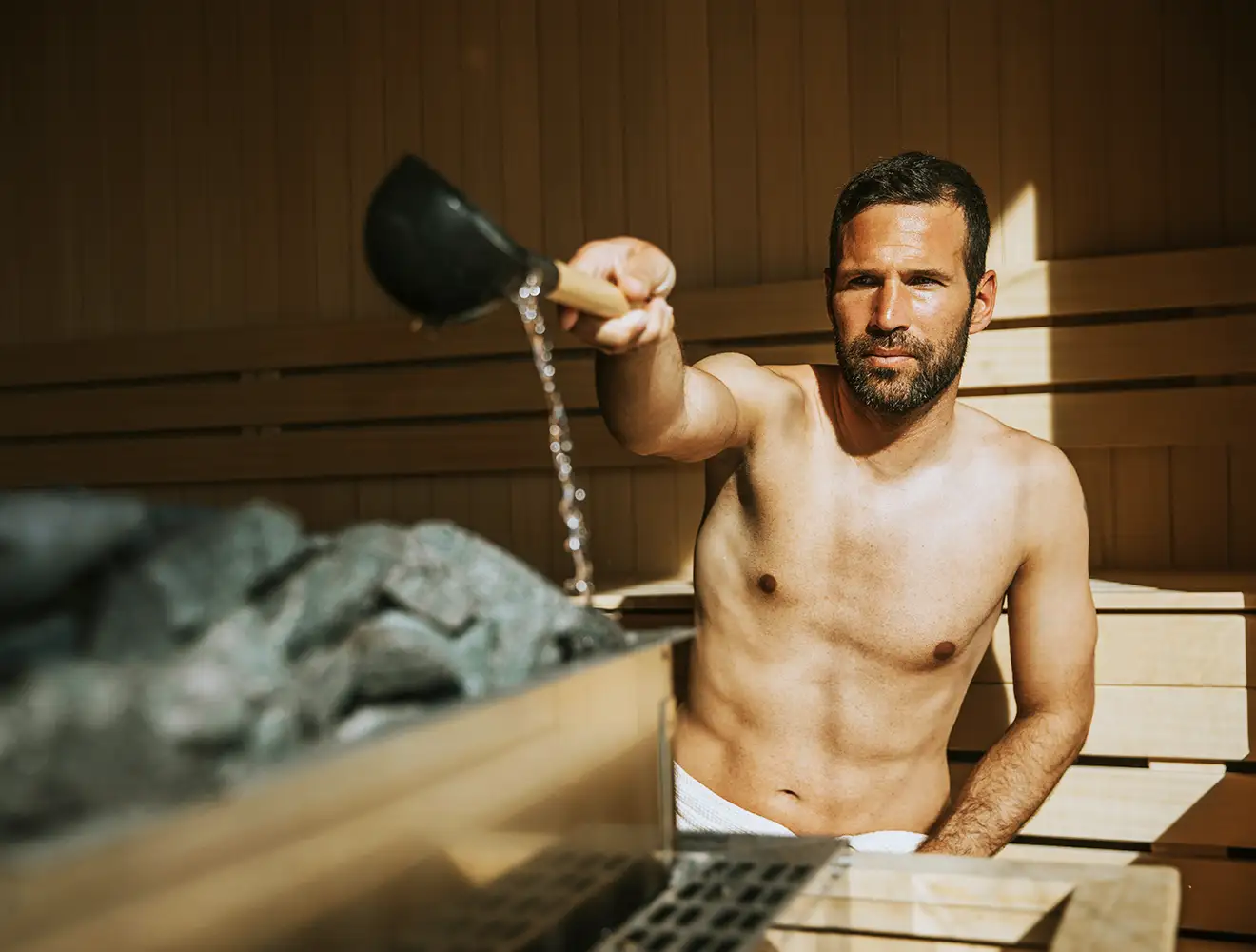 En mand hælder vand over varme sten i en træsauna til varmeterapi og longevity.