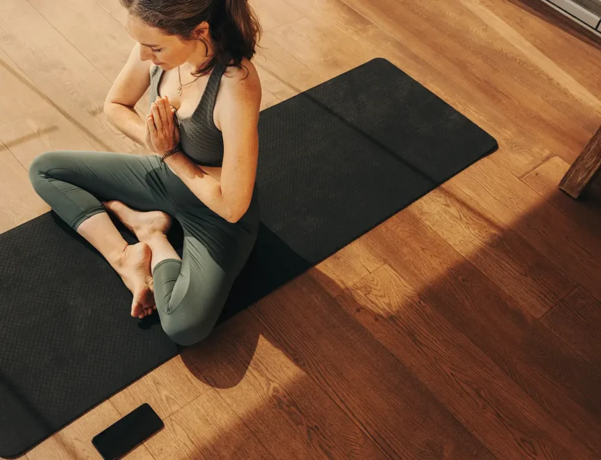 Woman meditating in a seated yoga pose on a mat on wooden floors.