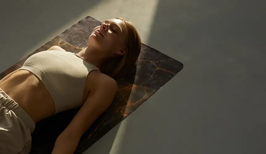 Woman lying peacefully on a yoga mat in soft sunlight, practicing relaxation or meditation during a calming wellness routine.