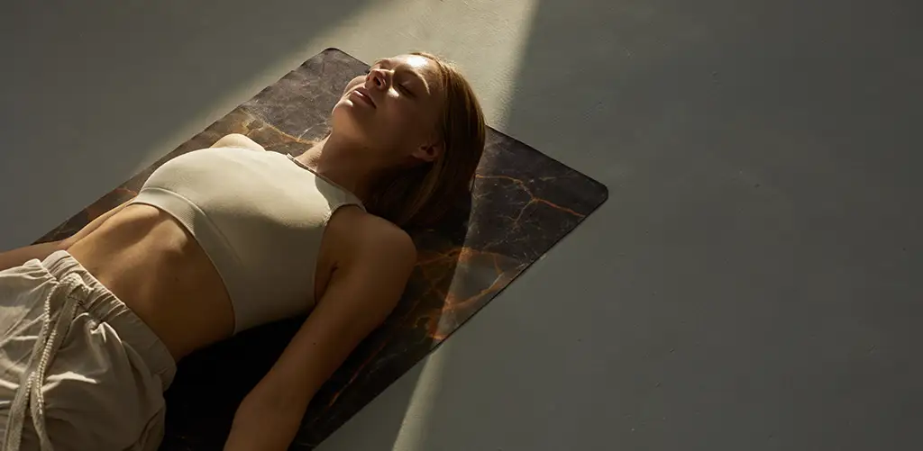 Woman lying peacefully on a yoga mat in soft sunlight, practicing relaxation or meditation during a calming wellness routine.
