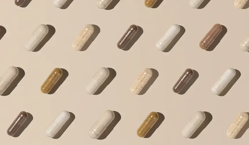 Cápsulas de suplementos dispuestas en un patrón ordenado y repetitivo sobre un fondo beige, en distintos tonos de blanco, tostado y marrón, que proyectan sombras suaves.
