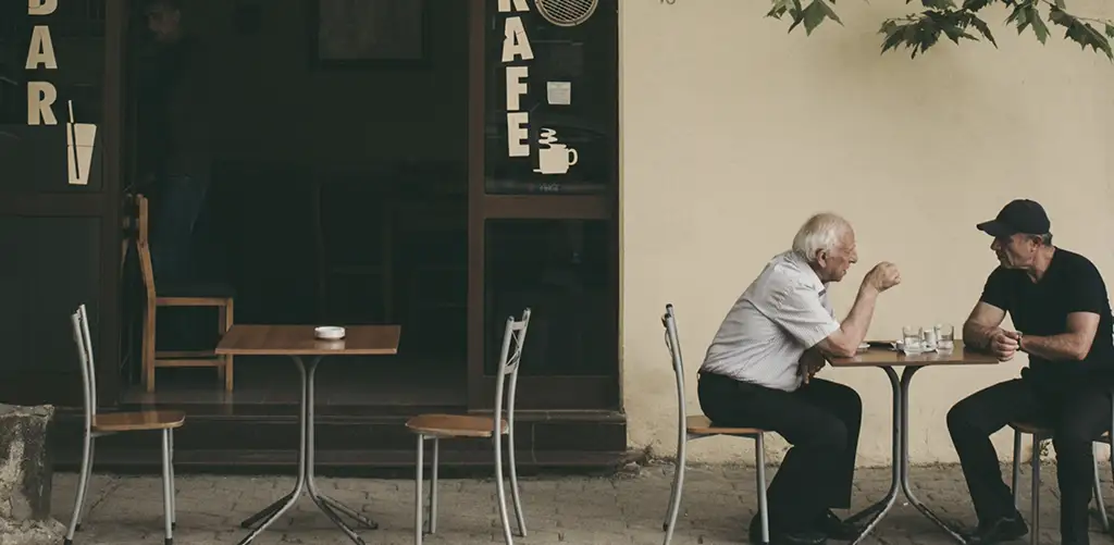 Due signori anziani seduti a un tavolino all'aperto di un bar, chiacchierano, mostrando come si possono avere legami sociali e compagnia anche quando si è più grandi.