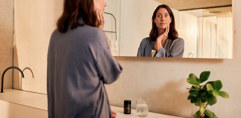 Una donna in bagno, che si tocca delicatamente il viso mentre si guarda allo specchio, con il prodotto Purovitalis Resveratol sul lavandino.