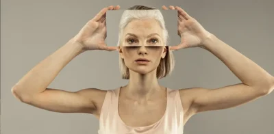 A woman holding a transparent panel in front of her face that shows an older version of her features, illustrating the contrast between youth and aging.