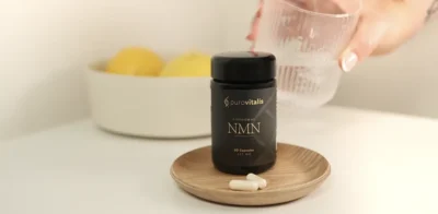 Flesje met Purovitalis Liposomal NMN-supplement op een klein houten bordje met capsules ervoor, op een witte tafel, met op de achtergrond een hand die een glas water vasthoudt.