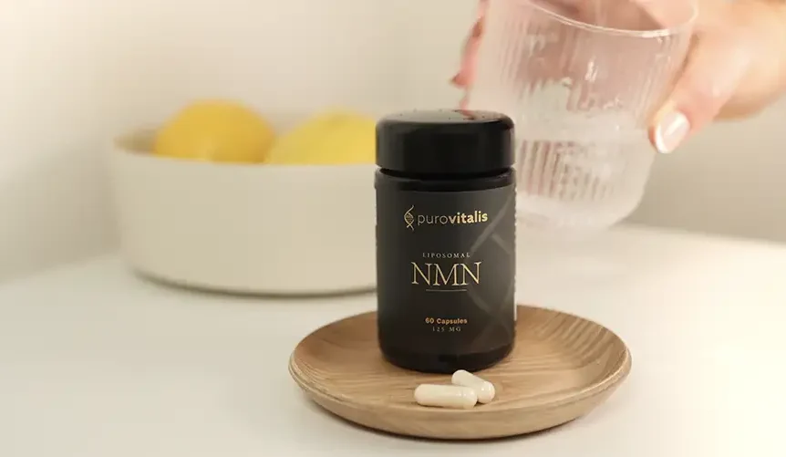 Un frasco del suplemento liposomal de NMN de Purovitalis sobre una pequeña bandeja de madera, con las cápsulas en primer plano, colocado sobre una mesa blanca, y una mano que sostiene un vaso de agua al fondo.