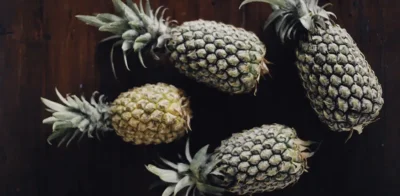 Five whole pineapples arranged on a dark wooden background, viewed from above, showing their textured green-brown skin and spiky leaves.