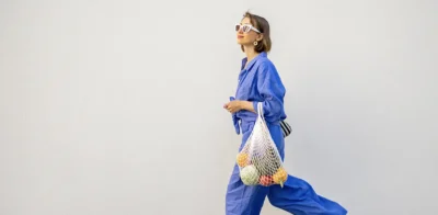 Woman in a blue outfit walking along a light-colored wall, carrying a reusable mesh bag filled with fresh fruits