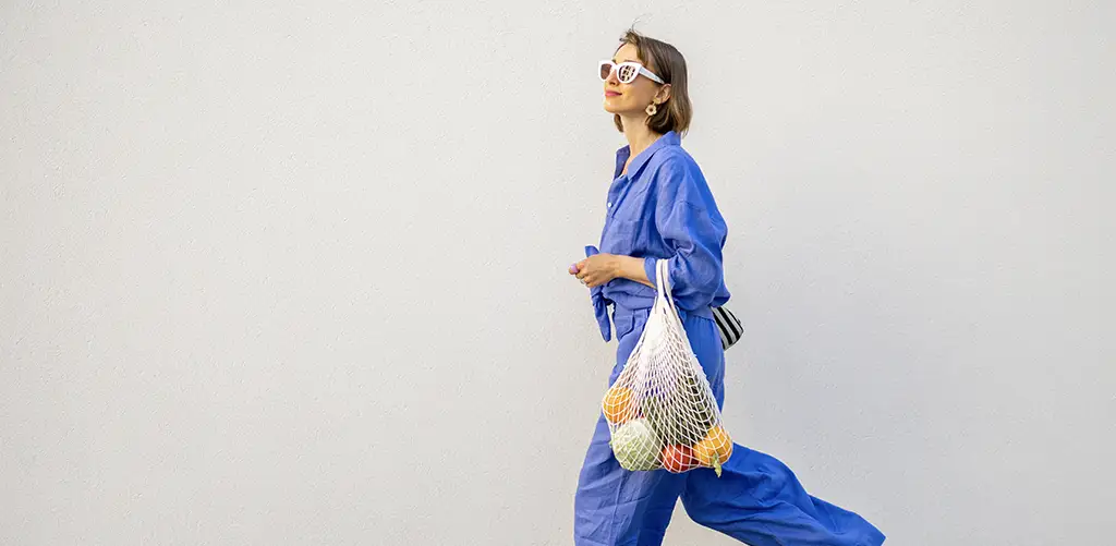 Woman in a blue outfit walking along a light-colored wall, carrying a reusable mesh bag filled with fresh fruits