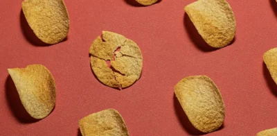 Een patroon van aardappelchips verspreid over een rode achtergrond, met in het midden één gebroken chip die eruit springt.