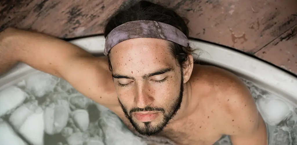 Ein bärtiger Mann mit einem Stirnband entspannt sich mit geschlossenen Augen in einem Eisbad, von oben gesehen.